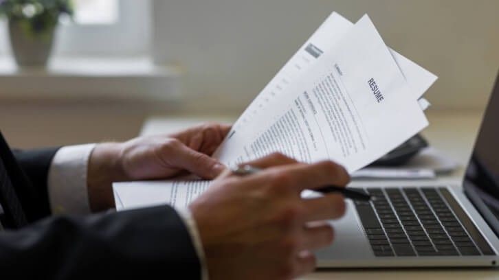 A person is holding papers and writing on a laptop.